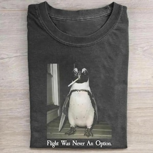 May include: A charcoal gray crew neck t-shirt featuring a graphic of a penguin holding a knife in its wing and a cigarette in its beak. Below the penguin, the text reads, "Flight Was Never an Option." The shirt has a short-sleeved design.