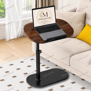 May include: A height-adjustable side table with a dark brown wood-grain finish oval top and a black metal base. A laptop is placed on the table surface, displaying a circular logo with the text MahnumCraft and Handmade with Love.