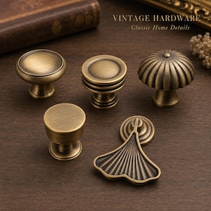 May include: Five antique brass cabinet knobs and pulls with varying shapes including round, ribbed, and fan-like designs. The text VINTAGE HARDWARE Classic Home Details is visible at the top. These metal hardware pieces feature a brushed finish.