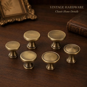 May include: Six round cabinet knobs with a brushed antique brass finish. Each knob features a flat circular top and a tapered pedestal base. The text VINTAGE HARDWARE Classic Home Details is visible in the upper right corner of the image.