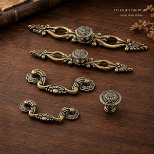 Peut inclure: Un ensemble de cinq pièces de quincaillerie pour meubles en métal couleur laiton antique, comprenant deux longues poignées ornées avec boutons centraux, deux petites poignées tombantes et un bouton rond, avec des détails floraux et volutes.