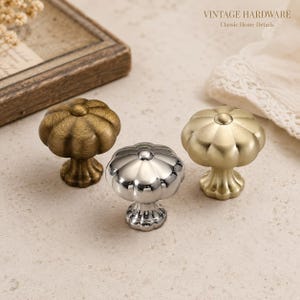 Op de afbeelding: Drie metalen kastknoppen met een pompoenvormig ontwerp en een geribbelde voet. De set bevat één knop met antieke messing afwerking, één met gepolijst chroom en één met geborsteld goud. De tekst VINTAGE HARDWARE Classic Home Details is zichtbaar.