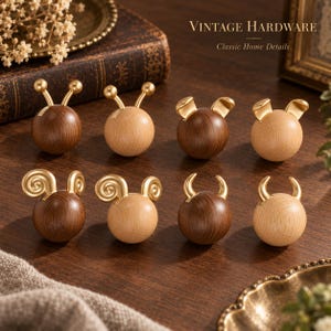 May include: Eight spherical cabinet knobs arranged in two rows, featuring dark walnut and light beech wood finishes. Each knob is topped with gold-toned metal accents in various shapes, including antennae, rounded ears, spiral horns, and curved horns.