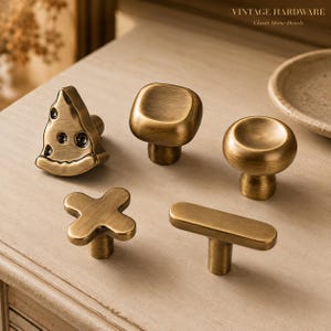 Peut inclure: Cinq boutons de placard en laiton avec une finition brossée. L'ensemble comprend une forme de part de pizza triangulaire, un carré arrondi, un bouton circulaire, une croix à quatre branches et une barre horizontale. Texte VINTAGE HARDWARE visible.