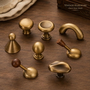 May include: A collection of eight brass cabinet knobs and pulls with varied shapes, including round, conical, arched, and angled designs. Some pieces feature dark wood accents. The text Vintage Hardware Classic Home Details is visible.