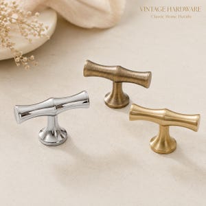 May include: Three T-shaped cabinet knobs featuring a bamboo-inspired design. One knob has a polished chrome finish, one has a brushed antique brass finish, and one has a satin gold finish. The text VINTAGE HARDWARE Classic Home Details is visible.