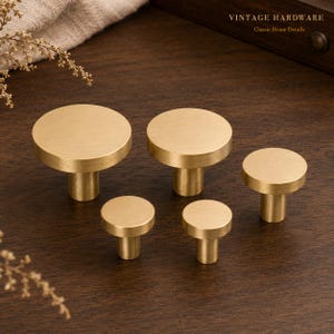 May include: Five circular cabinet knobs in a brushed gold finish, arranged in varying sizes. Each knob features a flat, round top surface and a cylindrical base. The text VINTAGE HARDWARE Classic Home Details is visible in the upper right corner.