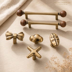 Op de afbeelding: Een collectie kastbeslag van messing, waaronder twee lange handgrepen met donkerhouten bolvormige uiteinden, een strikvormige knop, een wolkvormige knop, een koffieboonvormige knop en een X-vormige knop, allemaal met een matte antiek messing afwerking.