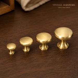 May include: Four round cabinet knobs in a brushed brass finish, arranged in a row from smallest to largest. Each knob features a smooth, domed top and a tapered base. The text VINTAGE HARDWARE Classic Home Details is visible in the top right corner.