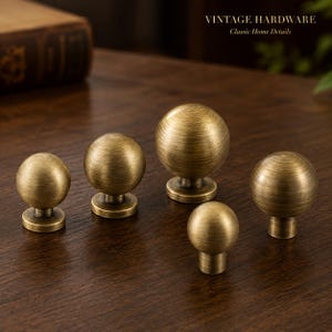 Op de afbeelding: Vijf bolvormige kastknoppen in een geborstelde antiek messing afwerking met een ronde voet. De knoppen variëren in grootte en hebben een getextureerd oppervlak met horizontale lijnen. De tekst VINTAGE HARDWARE Classic Home Details is rechtsboven zichtbaar.
