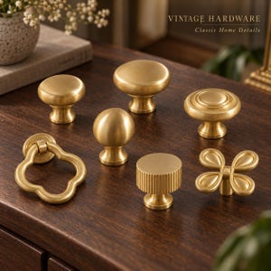 Puede incluir: Una colección de siete pomos y tiradores de armario de metal en tono latón con diversas formas, incluyendo diseños redondos, estriados y en forma de trébol. El texto VINTAGE HARDWARE Classic Home Details es visible en la esquina superior derecha.