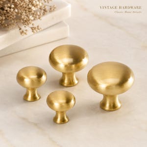 May include: Four round cabinet knobs with a brushed gold finish and a smooth, domed top surface. The knobs vary in size, featuring a flared base that tapers upwards to a circular head. The text VINTAGE HARDWARE Classic Home Details is visible.