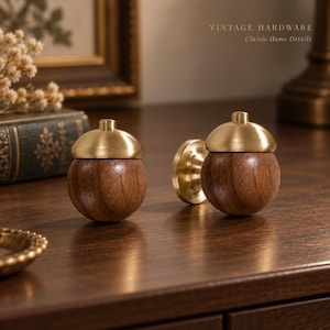 Puede incluir: Dos pomos de armario con forma de bellota que presentan una base esférica de madera de color marrón oscuro liso y una tapa de metal de latón pulido. El texto VINTAGE HARDWARE Classic Home Details es visible en la esquina superior derecha.