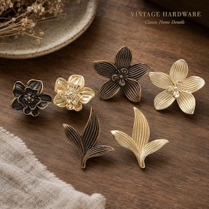 May include: A collection of six metal cabinet knobs in flower and leaf shapes, featuring dark bronze and polished gold finishes. The knobs include two five-petaled flowers, one layered flower, and two leaf-shaped designs. Text reads VINTAGE HARDWARE.