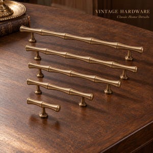 May include: Five brass cabinet pulls in varying lengths, each featuring a textured bamboo-inspired design with a matte finish. The pulls are arranged in a row, with the longest at the back and the shortest at the front. Text reads VINTAGE HARDWARE.