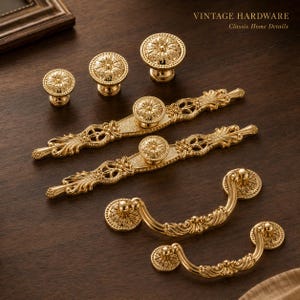 Puede incluir: Una colección de herrajes para armarios de metal dorado que incluye tres pomos redondos, dos tiradores rectangulares largos con detalles de filigrana ornamentados y dos tiradores colgantes curvos. El texto VINTAGE HARDWARE Classic Home Details es visible.