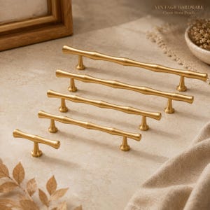 May include: A set of five gold-toned metal cabinet pulls with a textured bamboo-inspired design. The collection includes one single-knob pull and four bar pulls of varying lengths, each featuring a smooth, matte finish and two mounting posts.