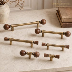 May include: A collection of six cabinet pulls in varying lengths, each featuring a brushed brass metal bar with dark brown wooden accents at the ends. The wooden ends are shaped as either spheres or cylinders, and each pull has two mounting posts.