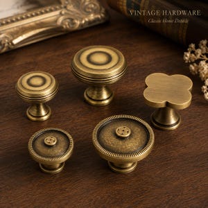 Peut inclure: Cinq boutons de placard en laiton avec une finition antique. Deux boutons sont circulaires avec des anneaux concentriques, un est en forme de trèfle, et deux sont circulaires avec un petit motif floral au centre. Le texte VINTAGE HARDWARE est visible.