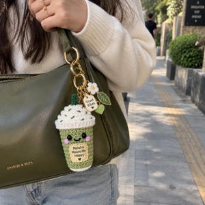 May include: A crochet matcha drink keyring attached to an olive green handbag. The keyring features a green cup with a white whipped top, a smiling face, and a tag that reads Matcha Makes Me Happy. It includes charms of a flower and green leaves.
