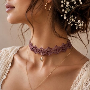 May include: A dark mauve crochet lace choker necklace featuring a repeating floral pattern and a central teardrop-shaped pendant with a white faux pearl. A thin gold-toned chain necklace with a small white bead hangs below the lace choker.