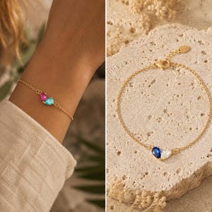 May include: A gold-toned chain bracelet featuring two pear-shaped gemstones. One version displays a pink and a light blue stone, while the other version showcases a deep blue and a clear white stone. Each bracelet includes a lobster claw clasp.