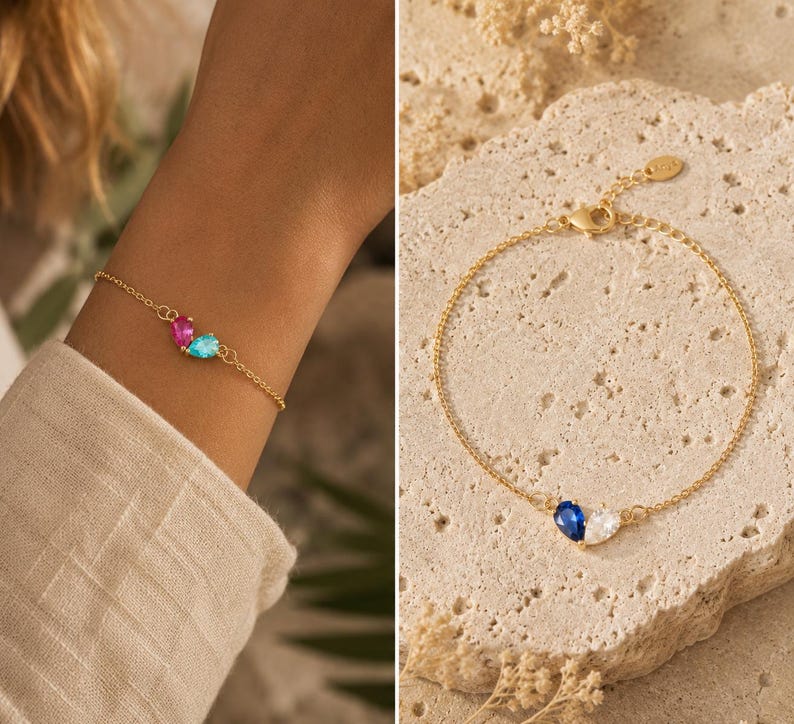 May include: A gold-toned chain bracelet featuring two pear-shaped gemstones. One version displays a pink and a light blue stone, while the other version showcases a deep blue and a clear white stone. Each bracelet includes a lobster claw clasp.