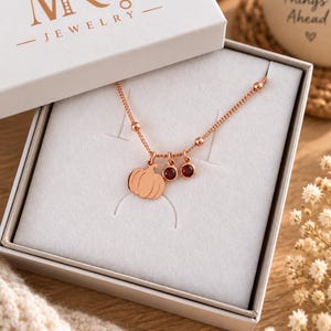 Peut inclure: Un collier couleur or rose avec un pendentif citrouille et deux petites breloques circulaires en pierre pr&eacute;cieuse rouge fonc&eacute;. Le collier poss&egrave;de une cha&icirc;ne d&eacute;licate avec de petites perles et est pr&eacute;sent&eacute; dans une bo&icirc;te cadeau blanche.
