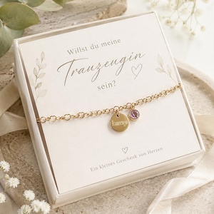 Puede incluir: Una pulsera de cadena dorada con eslabones en forma de corazón, que incluye un dije redondo grabado con la palabra Trauzeugin y una pequeña piedra circular morada, presentada en una caja blanca con el texto en alemán Willst du meine Trauzeugin sein?
