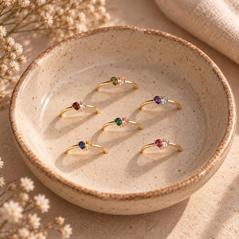 May include: Six gold rings, each featuring two small heart-shaped gemstones in different color combinations, arranged in a circular ceramic dish. The rings have thin bands and display various pairs of colored stones including red, blue, green, and pink.