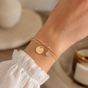Puede incluir: Una pulsera de doble cadena dorada con dos dijes delicados. Un dije es un disco circular dorado grabado con una ilustración de líneas de dos manos tocándose, y el segundo dije es una pequeña piedra de cristal redonda y transparente.