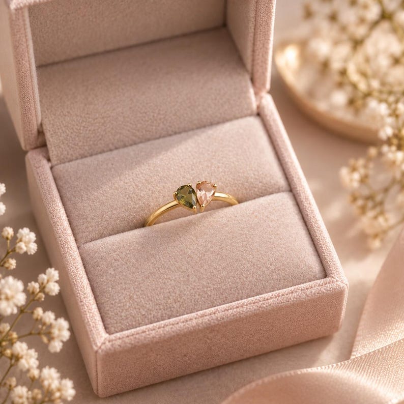 May include: A gold ring featuring two pear-shaped gemstones, one olive green and one pale peach, set side-by-side on a thin gold band. The ring is displayed inside a light pink velvet jewelry box with a soft, plush interior lining.