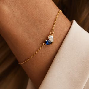 May include: A delicate gold chain bracelet featuring two pear-shaped gemstones arranged in a cluster. One gemstone is a deep blue sapphire color, and the other is a clear, colorless stone. The bracelet has a thin, polished gold-tone metal link chain.