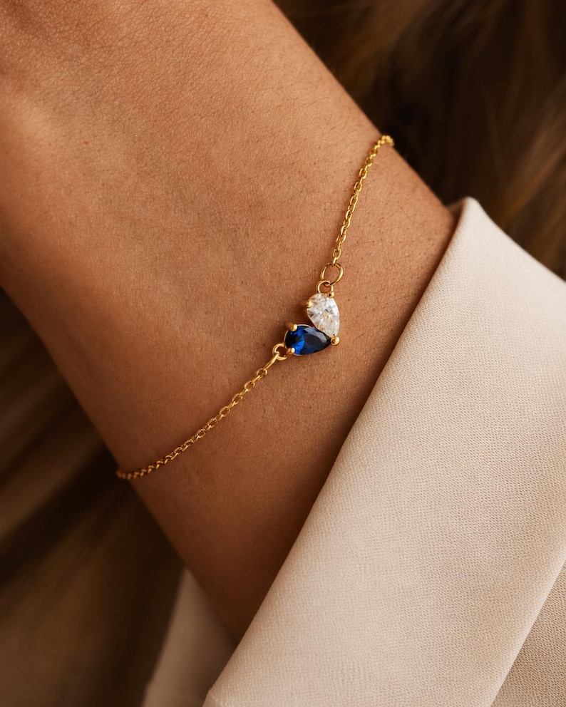 May include: A delicate gold chain bracelet featuring two pear-shaped gemstones arranged in a cluster. One gemstone is a deep blue sapphire color, and the other is a clear, colorless stone. The bracelet has a thin, polished gold-tone metal link chain.