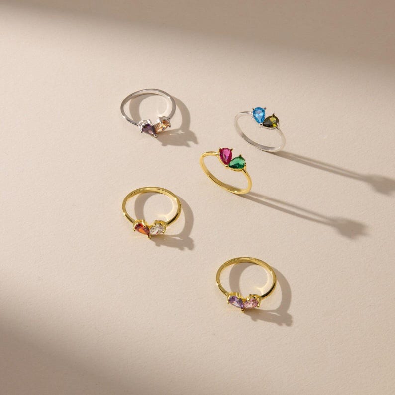 May include: A collection of five thin metal rings, each featuring two small teardrop-shaped gemstones in contrasting colors. The set includes silver-toned and gold-toned bands, with stones in shades of blue, green, pink, purple, and clear white.