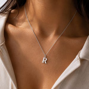 Könnte beinhalten: Eine silberfarbene Halskette mit einer feinen Kette und einem einzelnen Anhänger in Form des Großbuchstabens R. Der Anhänger hat eine glatte, polierte Oberfläche und hängt mittig an der dünnen Metallkette im Bereich des Schlüsselbeins.