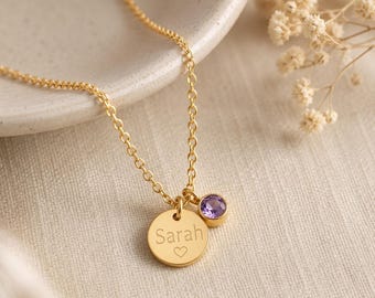 Op de afbeelding: Een goudkleurige ketting met een fijne schakelketting en twee bedels: een ronde hanger gegraveerd met de naam Sarah en een klein hartje, en een tweede bedel met een ronde, gefacetteerde paarse amethistkleurige steen.