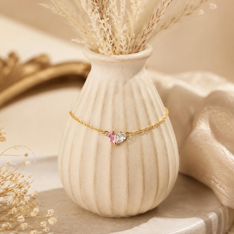 May include: A delicate gold chain bracelet featuring a central heart-shaped charm composed of two distinct stones, one light pink and one clear white, set in a polished gold-tone metal frame, resting against a ribbed, off-white ceramic vase.
