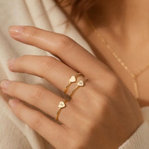 Könnte beinhalten: Drei goldfarbene Ringe mit kleinen herzförmigen Anhängern. Zwei Ringe werden am Ringfinger getragen, einer mit dem eingravierten Buchstaben S auf dem Herz und ein schlichter Herzring. Ein dritter Ring mit dem eingravierten Buchstaben R auf dem Herz wird am Mittelfinger getragen.