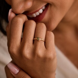 May include: A gold ring featuring a heart-shaped centerpiece composed of two distinct teardrop-cut gemstones, one in a deep olive green and the other in a soft peach tone, set on a thin, polished gold band that encircles the finger.