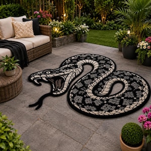 May include: A large, shaped area rug featuring a coiled snake design in black, grey, and cream. The rug depicts a snake with an open mouth, visible fangs, and a long tongue, with a textured, patterned body that follows the contours of the snake shape.