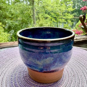 May include: A ceramic planter with a wide, circular opening and a tapered base. The upper portion features a glossy, deep blue and purple glaze with subtle color variations, while the lower section remains an unglazed, matte terracotta brown.