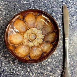 May include: A round ceramic bowl featuring a glossy brown and tan glaze with a radial, flower-like pattern radiating from the center. Beside the bowl lies a silver-toned metal dinner knife with an ornate, embossed handle design.