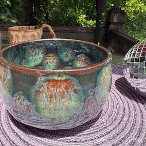 May include: A round ceramic bowl featuring a crystalline glaze with a blend of deep teal, forest green, and burnt orange circular patterns. The rim is a solid dark brown, and the interior displays a matching variegated color scheme.