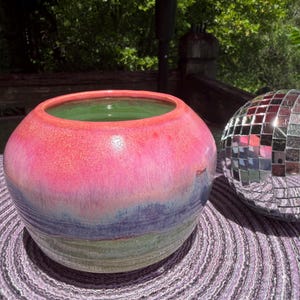 May include: A round ceramic bowl with a smooth, glossy glaze featuring a gradient of vibrant pink at the top, transitioning into shades of purple and blue, with a light green interior. The bowl has a wide opening and a rounded, bulbous shape.