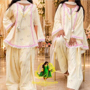 May include: A cream-colored tunic and loose-fitting trousers set featuring vertical yellow stripes and small floral patterns. The tunic has long sleeves, a V-neckline with pink and white lace trim, and decorative tassels. The set includes a matching scarf.