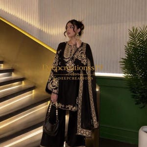 Op de afbeelding: Een zwarte driedelige set bestaande uit een tuniek met lange mouwen, een wijde broek en een bijpassende dupatta. De kledingstukken zijn voorzien van ingewikkeld goudkleurig borduurwerk langs de halslijn, mouwen en zomen. Een kleine zwarte handtas is inbegrepen.
