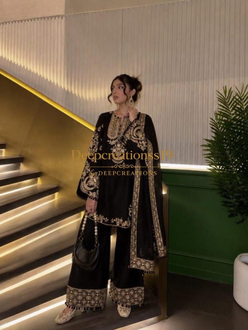Op de afbeelding: Een zwarte driedelige set bestaande uit een tuniek met lange mouwen, een wijde broek en een bijpassende dupatta. De kledingstukken zijn voorzien van ingewikkeld goudkleurig borduurwerk langs de halslijn, mouwen en zomen. Een kleine zwarte handtas is inbegrepen.
