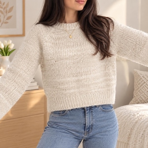 May include: A cream-coloured, long-sleeved knit jumper with a round neckline and ribbed cuffs and hem, paired with light blue denim jeans. The jumper features a textured, speckled yarn finish and a relaxed fit with raglan-style sleeves.