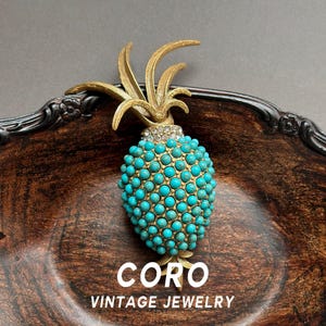 May include: A pineapple-shaped brooch featuring a textured gold-tone metal top and a body covered in small, round, turquoise-coloured beads. A band of clear, sparkling rhinestones separates the gold leaves from the beaded base. CORO VINTAGE JEWELRY.